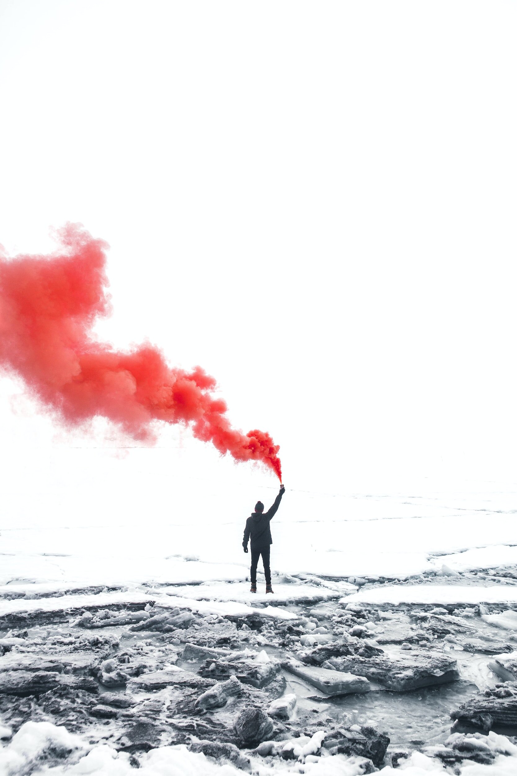 Person standing on a snowy icy landscape during winter, holding a red smoke flare emitting red smoke, with a blank or pale sky in the background.